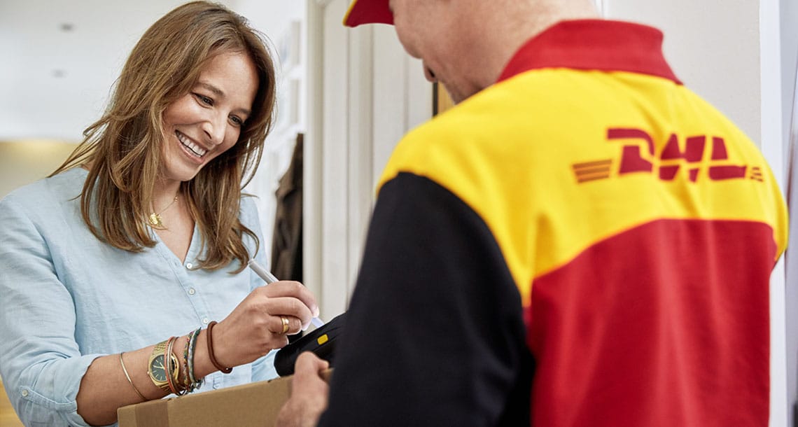 A woman at her door smiling and signing over an international delivery parcel from a DHL delivery man.